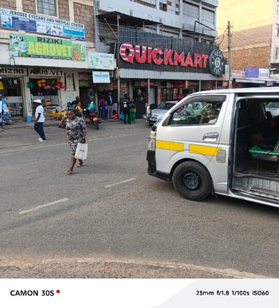 This image depicts a bustling daytime street scene in Machakos Central ward, Kenya. The weather appears clear with good visibility.
In the midground, a woman in a patterned dress, carrying a white shopping bag, walks across a tarmac road from left to right. To the right, a silver passenger van, likely a "matatu" common in Kenya, is parked or stopped with its side door open. It features distinctive yellow reflective strips along its side.
The background is dominated by a row of commercial buildings housing various businesses. A prominent red sign for "QUICKMART" supermarket is visible, along with a smaller "FRESH & EASY" sign. To the left of Quickmart, another building features signs for "JEROSEE EYECARE AND MEDICAL CENTER" and "RIZIKI AGROVET," which is displayed twice. Other visible shop signs include "FINECHEF," and partially obscured text like "BUY & SUPER" and "BUSINPC." Numerous people are scattered on the sidewalks, engaged in typical street activities like walking and standing near the shops. A motorbike is also visible near the storefronts. One person wears a black t-shirt with the number "80" on it.
The image metadata indicates it was captured with a CAMON 30S camera, at 23mm focal length, f/1.8 aperture, 1/100s shutter speed, and ISO60.