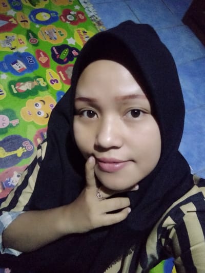 This image depicts a female individual in an indoor setting within the city of Lumajang, Indonesia. The subject, positioned centrally and looking directly at the camera, wears a black hijab and a light-colored striped shirt with black and tan vertical stripes. Her right hand is raised, with the index finger touching her lower lip or chin area; a ring with a transparent stone is visible on her right ring finger. The background on the left features a vibrant green mat adorned with multicolored cartoon characters, shapes, and numbers. To the right, a plain, muted blue surface is visible. The scene is illuminated by frontal lighting.