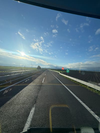 The image displays a forward view from inside a vehicle on an asphalt highway in Carapelle, Italy. The road is a two-lane thoroughfare with white solid and dashed lane markings, extending into the distant horizon. To the left of the primary road, a parallel service road or track is visible, separated by a metal guardrail. Beyond this, flat fields and multiple wind turbines are present under a bright sky. To the right of the main road, a metal guardrail separates the highway from a tilled agricultural field. Further right, a red rectangular sign with white text (possibly "SOS") and a green rectangular sign with a white right-pointing arrow are affixed to posts. Numerous wind turbines are visible in the distant landscape on both sides of the road. The sky is blue with scattered white clouds, and the sun, positioned in the upper left, creates a bright lens flare. No individuals or active interactions are visible.