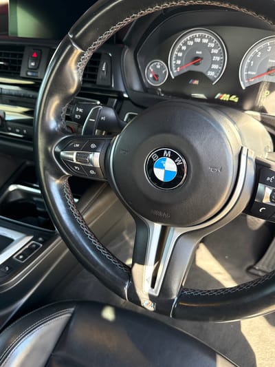 The image captures the interior of a BMW M4 from the driver's perspective, focusing on the steering wheel and instrument cluster. The scene takes place during daylight, with bright sunlight casting reflections within the car's cabin.
The central subject is a black leather multi-function steering wheel, detailed with red and blue stitching, a signature of BMW M-Sport design. The iconic BMW logo is centrally positioned, with "AIRBAG" visible below it. The left side of the steering wheel features control buttons, including "LIM" (Limiter) and "RES" (Resume) for cruise control, while the right side has "M1" and "M2" buttons for customizable drive modes. A small M logo is present on the lower spoke.
Behind the steering wheel, the instrument cluster is visible, showing a tachometer on the left marked "x 1000 1/min" with a "READY" indicator, and a speedometer in the center displaying "km/h" up to "330". Both needles rest at zero, suggesting the car is stationary. A smaller gauge is located beneath the speedometer. The "M4" logo is distinctly visible on the tachometer, and another tri-color M logo appears near the speedometer.
Additional details include parts of the black leather car seat with white stitching, interior air vents, and various buttons on the dashboard and door panel. The overall setting is the driver's cockpit of a performance vehicle. The location context for this scene is Blouberg, South Africa.