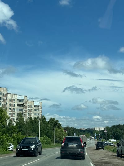 This is a candid, eye-level, medium shot of a four-lane road in a suburban area of Priogorodniy, Russia. The sky is a vibrant blue with scattered white clouds. In the foreground, a black SUV is driving in the left lane, its license plate reading "Y044YP 63". To the left of the road, a multi-story, Soviet-era apartment building is visible, with trees lining the street in front of it. Further down the road, several other vehicles, including a white SUV, a truck, and other cars, are driving in the same direction. The scene appears to be captured from another vehicle, suggesting a snapshot of daily life or travel in this Russian city.