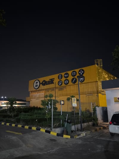 At night in Kelapa Dua, Indonesia, a low-angle shot shows the yellow B-Quik store. The building has several icons on its facade. There is a parking sign and a wire fence along the perimeter of the building. The pavement is painted with yellow and black stripes. A car is partially visible parked beside the building. Vegetation is growing around the fence and there are streetlights illuminating the area. The sky is dark.