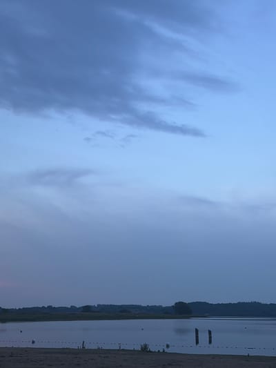 The image depicts a serene lake scene in Preetz, Germany, likely at dusk or dawn given the soft, diffused light and the muted colors. The sky dominates the upper portion of the frame, transitioning from a deep, dusky blue at the top to a lighter, paler blue towards the horizon. Wisps of clouds, appearing as subtle smudges of darker blue and grey, are scattered across the sky, adding texture and depth.

The middle ground features a tranquil body of water, reflecting the pale sky. The surface of the lake is mostly still, with only gentle ripples visible. Several white buoys are scattered across the water, marking out a designated area. A rope with red and white markers connects some of these buoys, further delineating a section of the lake. Two dark wooden posts rise from the water near the right side of the frame, their purpose unclear but adding a touch of rustic structure to the scene.

The foreground consists of a sandy beach or shoreline. The sand appears damp and slightly textured. Beyond the lake, the horizon is lined with a dark, silhouetted band of trees and low hills, creating a natural backdrop that merges with the sky. The overall impression is one of quietude and natural beauty. There are no people or animals visible in the frame. The environment is peaceful and undisturbed, suggesting a natural setting within or near the city of Preetz.