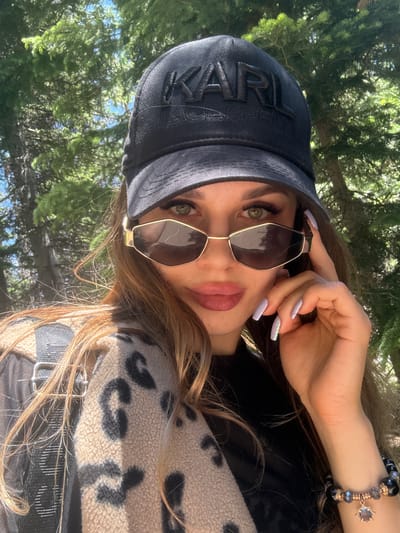 A woman with long brown hair and green eyes is taking a selfie outdoors. She is wearing a black baseball cap with a raised logo, stylish sunglasses with gold frames, and a beige fleece jacket with a black leopard print. A beaded bracelet adorns her left wrist. She is touching the side of her sunglasses with her right hand. In the background, lush green trees suggest a natural setting, possibly a park or forest. The image appears to have been taken during daylight hours with bright sunlight filtering through the leaves. The precise location in Allenspark, United States, is not identifiable from the visual cues alone.