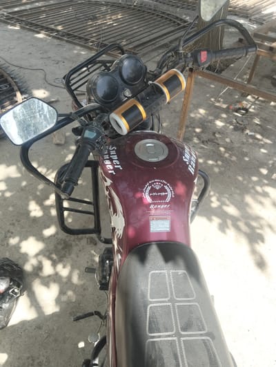 A close-up, overhead shot showcases the front portion of a maroon motorcycle parked outdoors. The motorcycle features a black handlebar with a red switch on the right grip and a silver mirror on the left. A cylindrical speaker with gold accents is attached to the handlebar. The motorcycle's fuel tank displays decals including "Super Ranger" in white lettering and a circular emblem with text in what appears to be Arabic script. A black seat is visible behind the fuel tank. The ground beneath the motorcycle is composed of dirt and concrete, with shadows of tree branches cast upon it. In the background, a metal structure, possibly a fence or gate under construction, is present. To the left of the motorcycle, a black plastic bag containing debris lies on the ground. The scene suggests a location in the city of Danghara, Tajikistan, possibly a workshop or storage area.