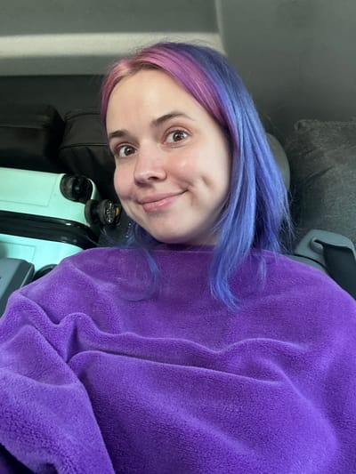 The image is a selfie of a person with purple and blue hair. The person is wearing a purple, fleecy top and has brown eyes. A light blue suitcase is visible in the foreground. The background shows parts of a gray couch. No location in Villeneuve-Loubet, France, can be determined from this image.