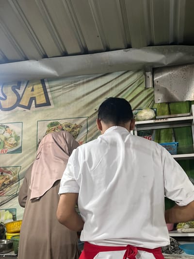 The image depicts the rear view of two individuals at a food stall in Boja, Indonesia. One person, wearing a white chef's coat with a red sash, stands facing away from the camera. The other individual, to the left, is covered in a beige hijab and a brown abaya, also facing away. Behind them, a banner with the word "SA" in yellow letters is visible. Shelving units with green banana leaves and a white cabbage are also present. The environment appears to be an outdoor or semi-outdoor setting with a corrugated metal roof.