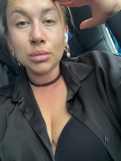 A woman with blonde hair and light skin is captured in a close-up, overhead shot, seated inside what appears to be a vehicle. She is wearing a black satin button-up shirt with the collar open, revealing a black ribbed bra underneath. The woman has on a black mesh choker necklace and gold hoop earrings, one of which features a small pink bead. A white wireless earbud is visible in her left ear. Her right hand is raised to her forehead, with her fingers splayed. The background consists of dark leather car seats. The image does not provide any specific information about the location in the city of Подберезье, Russia.