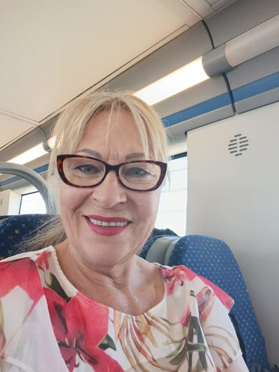 A blonde woman wearing red-rimmed glasses and a floral print top smiles for a selfie on what appears to be a train. She has bright pink lipstick on and her hair is pulled back. The background shows the interior of a train carriage, with blue patterned seats visible and overhead fluorescent lighting. The image has a bright, casual feel. There is no indication of the city of Palmela, Portugal, in the image.