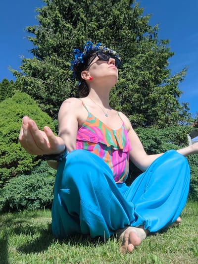 A person with dark hair, adorned with a blue floral crown and sunglasses, meditates outdoors in a cross-legged pose. They are wearing a vibrant, multi-colored tank top with ruffles and loose, bright blue pants. Their hands are resting in their lap, with the right hand in a mudra. The person's head is tilted back, and their eyes are closed, suggesting a state of calm or focus.

The setting is a grassy lawn with lush green bushes and trees in the background. The sky is a clear, bright blue, indicating a sunny day, likely in the late morning or early afternoon due to the shadows cast on the grass. The overall atmosphere is peaceful and serene, hinting at a practice of mindfulness or relaxation. The location context of Dzērumi, Latvia, suggests a natural, possibly rural or suburban outdoor environment. There is no visible text in the image.
