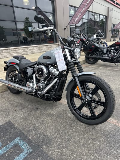 This is a medium-shot, eye-level, full-color photograph of a gray Harley-Davidson motorcycle parked on an asphalt surface outside a dealership. The motorcycle is the primary subject and is angled slightly towards the viewer's left. It is a street bike with a black seat, black wheels with silver spokes, and chrome accents on the engine and exhaust. A price tag is attached to the handlebars.
In the background, a building with large windows and a stone facade is visible. A banner with the text "BARNES HARLEY-DAVIDSON" is hanging from the building. Other motorcycles are parked in front of the building, some of which are black. The sky is overcast, suggesting a cloudy day. The ground in the foreground has faint blue markings that appear to be part of a parking lot designation. The scene is set in a commercial area, likely a motorcycle dealership in the Township of Langley, Canada.