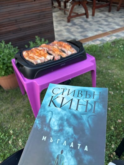 This image captures an outdoor leisure scene in Polena, Bulgaria. In the immediate foreground, a book titled "СТИВЪН КИНГ" (Stephen King) and "МЪГЛАТА" (The Mist) in Bulgarian is held by an unseen individual, angled towards the viewer. The book cover features a blue-grey, misty design with a silhouetted figure facing away, standing amidst vague, tentacle-like forms.
Beyond the book, a bright magenta-purple plastic stool or low table supports a black electric grill. On the grill, approximately four to five pieces of raw or partially cooked meat, possibly pork chops or ribs, are laid out on the grates, displaying a reddish-orange seasoning.
The ground surrounding the setup is covered in green grass interspersed with small white clover blossoms. To the left of the purple table, a brown terracotta-style pot holds a leafy green plant. In the background, a dark brown wooden structure or fence with horizontal planks is visible on the left. To the right, a paved patio area with grey square tiles in a geometric pattern extends, with blurred wooden outdoor furniture (tables and chairs/benches) further back. The overall lighting suggests natural daylight.