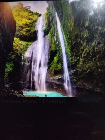 This is a photograph of a tall waterfall cascading down a rocky, lush green cliffside. The water plunges into a bright turquoise pool at the base of the falls. A solitary figure, appearing as a small silhouette, stands near the edge of the water, looking towards the waterfall. The surrounding environment is densely vegetated with vibrant green foliage covering the cliffs. Text is visible at the bottom of the image, reading "Indonesia, Mana Pilihanmu?....". The scene depicts a natural landscape, likely a tourist attraction, given the text and the clear view of the waterfall.