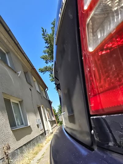 The image depicts a close-up, low-angle view of the rear of a dark-colored vehicle, primarily focusing on the taillight and a portion of the car's body. The taillight is red and appears to be a modern design with internal reflective elements. The vehicle's dark exterior shows some signs of wear and dust.

To the left of the vehicle, a light-colored, textured building is visible, appearing to be an apartment or residential building. It has several windows with white frames and a gutter system along the roofline. The building's facade is rough and weathered. A narrow path or sidewalk runs along the base of the building, with dry grass and some weeds growing alongside it. Above the building, a bright blue sky is visible, with the branches of a green coniferous tree extending into the frame from behind the vehicle.

The overall scene suggests an outdoor urban or suburban environment on a clear day, with a focus on the juxtaposition of the vehicle and the adjacent building. The vantage point is very low, looking up towards the sky and the building. The location is identified as Kostelec na Hané, Czechia.