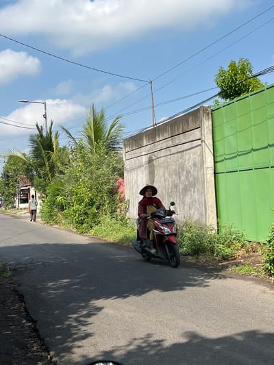 A person wearing a conical hat and a red scarf rides a red scooter down a paved road. They are on the right side of the road, with the scooter facing towards the viewer. The road is lined with lush green foliage and a tall, gray concrete wall with a vibrant green metal fence attached. In the background, palm trees and more greenery are visible, along with a street lamp. To the left of the road, a man walks away from the viewer, also on the right side of the road. The sky above is a clear blue with scattered white clouds. This scene appears to be in a rural or suburban area in Jati Alun Alun, East Java.