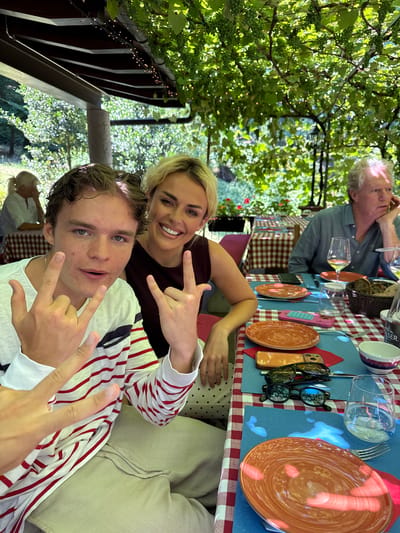 A medium shot captures three individuals at an outdoor table with a checkered tablecloth. The table is set with orange plates, glasses, and a basket of bread. Overhead, grapevines with green grapes cast dappled light on the scene. In the foreground, a young man with curly brown hair grins at the camera, holding up his hands in a rock-and-roll gesture. He wears a striped shirt and light-colored pants. To his right, a woman with blonde hair smiles broadly, also making a similar hand gesture. Further back, an older man with gray hair rests his chin on his hand, looking to the side. The location is Agno, Switzerland.