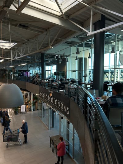 This image depicts the interior of a multi-level commercial building, possibly a market or shopping center, located in Eschau, France. The space features high ceilings with exposed light-colored structural beams, trusses, and multiple skylights, admitting natural light. Large dark support columns punctuate the open area, and extensive glass paneling forms exterior walls, providing views of an indistinct outdoor environment.
A prominent, dark-railed, curved mezzanine occupies the mid-ground, serving as the location for a restaurant identified by a dark banner displaying "AUX PETITS OIGNONS" in white lettering. Numerous tables with dark chairs are arranged on this upper level, some featuring red tablecloths, water bottles, bread baskets, and wine glasses. At least two individuals are present on the mezzanine: one male in a blue plaid shirt is seated with his back to the viewer in the right foreground, and another person is seated further to the left.
The lower level features light-grey tiled flooring. A large, grey, bell-shaped pendant light fixture hangs from the ceiling in the left foreground. Several individuals are present on this floor: an elderly man in a dark blue jacket pushes a silver shopping cart containing items, moving towards the left. An elderly woman in a pink jacket stands near an empty dark wooden table with two chairs, positioned below the mezzanine. Other individuals are visible walking or standing further in the background. Retail displays featuring plants and decorative items are arranged along the ground level, alongside a white freestanding sign. Glass storefronts line the base of the mezzanine, defining additional commercial spaces.