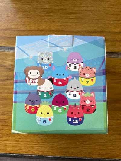 The image shows a close-up of a box with a colorful design featuring cartoon characters on a grassy field, likely a soccer field, with stadium seating in the background. The characters are various stylized animals and fruits, each wearing a soccer jersey with a number. They are arranged in rows. From left to right, top row: a blue bat with number 4, a purple seal with number 3, a pink leopard with number 7, and a white and blue spotted bear with number 10. Middle row: a brown Bigfoot-like creature with number 11, a gray shark with number 8, and a white cat with number 1. Bottom row: a purple creature with number 9, a yellow pineapple with number 5, a pink strawberry character with number 2, and a purple character with number 6. The box is placed on a wooden surface. The image does not contain any recognizable landmarks or elements that would suggest a specific location in Platja D'aro, Spain.