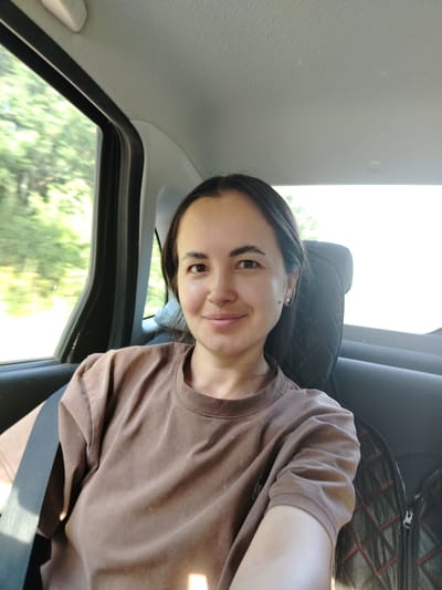 A young woman with dark hair and a nose ring is taking a selfie inside a vehicle. She is wearing a brown t-shirt and a seatbelt. Trees and foliage are visible through the window, suggesting a rural or suburban setting. The interior of the car shows a headrest and part of a seat.