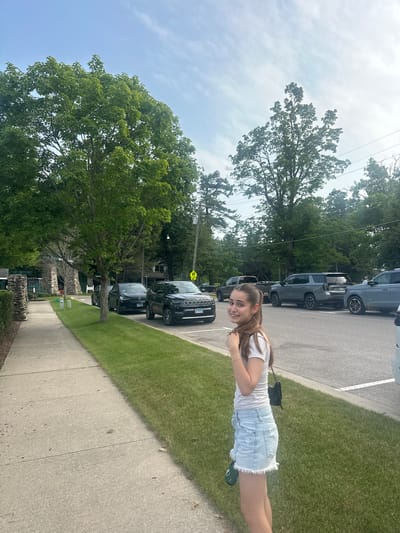 A young woman with long brown hair, wearing a white t-shirt and light blue denim shorts, stands on the grass next to a sidewalk in Harbor Springs, Michigan. She is smiling at the camera and holding her hair. Behind her, cars are parked along a street, and mature trees with green foliage line the sidewalk and parking area. The sky is partly cloudy with a soft, natural light. In the background, there appears to be a stone structure and some buildings, suggesting a park or a well-maintained street.