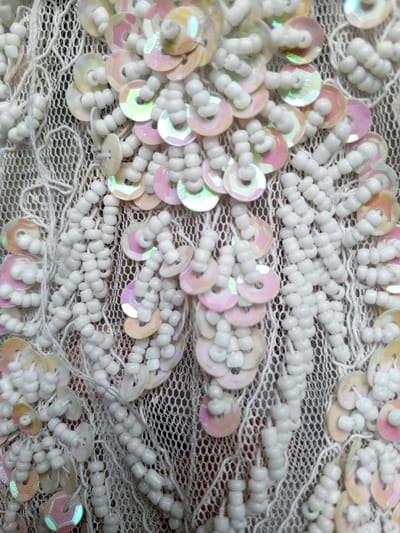 This close-up image from Houaykhang, Laos, showcases an intricately embellished textile. The primary subjects are numerous small, opaque white or off-white seed beads and iridescent sequins meticulously sewn onto a delicate, light-colored lace or mesh fabric.
The beads are arranged in dense clusters and elongated, organic patterns, forming textured, raised designs that resemble floral or abstract motifs. These beaded sections cover much of the underlying fabric. Interspersed among and within the beadwork are numerous small, circular sequins. These sequins display an iridescent quality, shifting in color to reveal soft hues of pink, peach, pale green, and yellow depending on the angle of light. They are often grouped in clusters or along the edges of the beaded patterns, sometimes attached by small strings of beads.
The background consists of a fine, open-weave lace, visible where the bead and sequin work is less dense, adding a subtle, delicate texture beneath the embellishments. The image captures the result of a detailed crafting or embroidery activity. The lighting is soft, highlighting the reflective properties of the sequins and the tactile quality of the beads. There is no visible text. The overall scene suggests a decorative fabric, possibly for apparel or home decor, characterized by its elaborate handiwork and shimmering details.