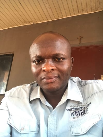 The image depicts a scene in the city of Egboje, Nigeria, showcasing a close-up portrait of an adult male. The individual, dark-skinned and bald, gazes directly at the camera with a neutral expression. He wears a light blue long-sleeved button-up shirt detailed with dark stitching, chest pockets, and text/graphics on the right side, including "AUTHENTIC DEPARTMENT" and "NINETEEN SIXTY SEVEN." The background comprises a textured light brown wall. To the subject's left, a dark rectangular opening, possibly a window, exhibits a reflection. To his right, a reddish-brown door or wall section is visible, with a small, light-colored crucifix mounted on the wall above it. The upper portion of the frame shows a paneled ceiling, likely wood or PVC, indicating a sheltered outdoor or semi-indoor environment such as a porch or veranda.