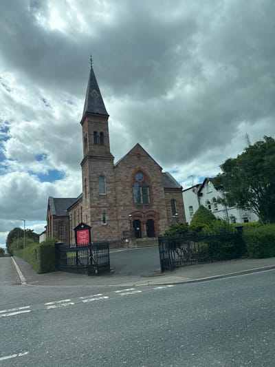 The image shows a stone church with a tall, pointed steeple on a cloudy day. The church is located in Belfast City, United Kingdom. A red sign in front of the church reads "KNOCK PRESBYTERIAN CHURCH. SUNDAY SERVICE 11.00 AM. REV. ROBERT HANNIN M.THEOL." A black wrought-iron fence surrounds a portion of the church grounds. To the right of the church, there is a white building with windows. The foreground of the image is a road with white double lines painted on it. Green hedges and trees are visible along the side of the road and around the church. The sky is filled with dramatic, grey clouds with patches of blue sky visible.