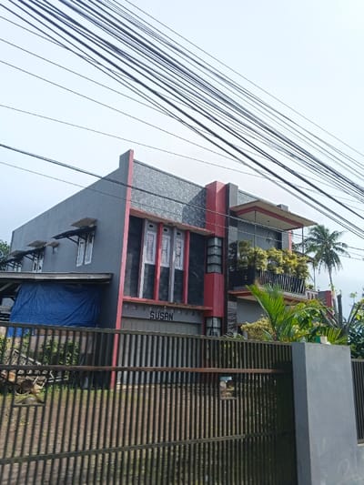 The image presents a two-story modern house, primarily gray with distinct red vertical architectural elements, situated under a bright, clear sky. A dense network of black electrical and utility wires stretches across the upper portion of the frame.
The house features a dark gray textured facade on its upper level and smooth lighter gray walls on the lower. On the ground floor, a roll-up garage door has the name "SUSAN" clearly visible in white letters above it. Multiple white-framed windows are distributed across both floors, some equipped with small awnings. To the left side of the house, a blue tarp covers an unidentified object beneath a lean-to structure. On the right, a second-floor balcony is adorned with vibrant green and yellow-leafed plants. Tall palm trees are visible in the background, behind the house on the right.
The property is enclosed by a dark metal fence with vertical bars, partially obscuring the lower section of the house and the front yard. The bright, clear sky suggests it is daytime with sunny weather. No people or specific activities are depicted in the scene, which appears to be a static view of a residential property. The architectural style, prominent overhead wires, and tropical vegetation are characteristic of residential areas, consistent with locations like Carcar, Philippines.