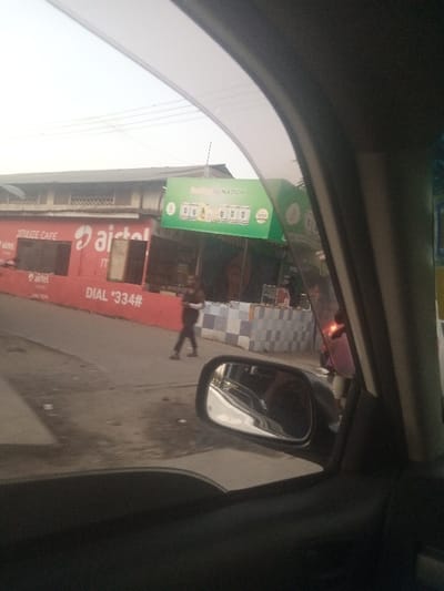 From inside a car, an Airtel-branded storefront is visible across the street. The building is painted red and advertises "DIAL *334#". Adjacent to it, a smaller shop with a green sign displays "Smile Wi-Fi NATION". A person is walking across the street in front of the shops, and another individual is partially visible near the second shop. The car's side mirror is in view, reflecting the interior of the car. The sky is overcast.