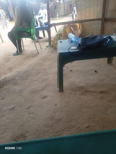 The scene depicts an outdoor or semi-sheltered area in Ushapa, Nigeria, characterized by an uneven dirt ground and visible wooden structural elements. In the mid-ground, a person with long dark hair, wearing a light-colored top and dark trousers, is seated facing away from the camera on a bright green plastic chair. Light blue sandals are visible on their feet. To their left and further back, another person with a bald head and a white shirt is seated at a blue plastic table containing several brown bottles and a white object. Blurry figures are present in the distant background, including one person in a patterned dress walking on the far left. In the foreground, a dark green plastic table holds a clear blue plastic water bottle lying on its side and a black shoulder bag next to a dark circular object. Behind this table, a large crumpled light brown sack or tarp rests against a wall with light-colored square tiles featuring a repetitive pattern. The area is illuminated by bright daylight, suggesting an open-air gathering or casual setting.