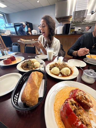 A woman is seated at a table in a restaurant, appearing to enjoy a meal. Various plates of food are arranged on the table, including stuffed peppers in sauce, meat with mashed potatoes, and a bread basket containing a loaf of bread. Other people are visible in the background, indicative of a dining establishment setting. The location is in Des Plaines, United States of America.