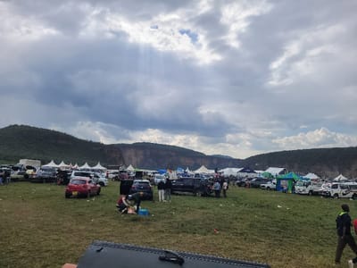 The image displays an outdoor gathering in Olkaria ward, Kenya, situated within a large, open grassy field. The background features a landscape of densely vegetated hills and prominent, steep rocky cliffs under a heavily overcast sky, with distinct crepuscular rays penetrating the clouds in the upper left. Numerous vehicles, including sedans, SUVs, and vans of various colors, are parked across the field, many with open trunks or doors. A multitude of white and colored canopy tents are erected throughout the mid-ground and background, indicating an event setup. Multiple individuals are present, either walking or standing in groups near the parked vehicles and tents. In the mid-ground, two individuals are observed kneeling on the grass next to a dark SUV with its trunk open, attending to items spread on a mat. Scattered debris is visible on the ground. The foreground contains the partial view of a dark, angular object.