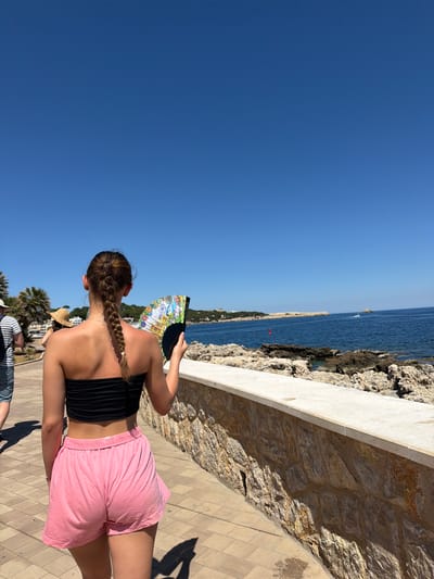 A woman is photographed from the back, holding a colorful fan in front of her face. She has her hair braided, wears a black top, and pink shorts. The scene is set along a stone pathway next to a rocky coastline, with the sea visible in the background. The sky is clear and blue. Based on the coastal scenery and architectural style, the location appears to be Cala Rajada, Spain. Other people are visible in the background.