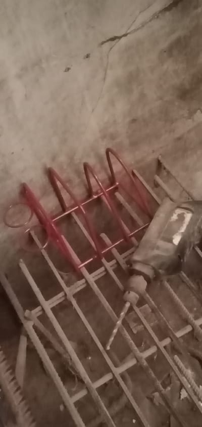 This is a close-up, low-angle, indoor shot of construction materials in what appears to be a workshop or storage area in Sumput, Indonesia. In the foreground, a metal reinforcement grid, likely for concrete, is visible, with rebar rods laid out in a crisscross pattern. A red metal object, possibly a tool holder or part of a structure, with several U-shaped loops, is placed on top of the grid. To the right of this, a black and silver power tool, likely a drill or hammer drill, is positioned on the rebar, with its chuck and a drill bit extending downwards. The background consists of a textured, greyish wall, possibly concrete, with some subtle cracks and variations in tone. The lighting is dim, casting soft shadows and contributing to a muted, earthy color palette. No people are visible in the frame.