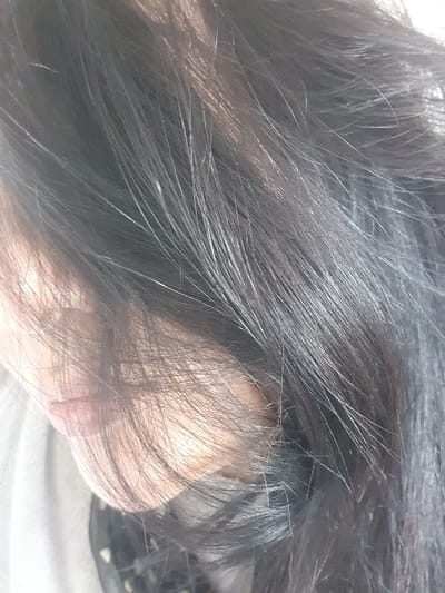 The image displays a close-up view of a subject's head, primarily focusing on dark, straight hair that heavily obscures the left portion of the face. Approximately two-thirds of the frame, from the top right extending downwards, is filled with the subject's hair, exhibiting individual strands and some light reflections. A segment of the subject's light-toned skin is visible on the left side, including a portion of the upper lip and the curve of a cheek. A light-colored garment, possibly gray or off-white, is discernable beneath the chin area on the far left. A darker, possibly patterned fabric is partially visible beneath the hair near the bottom center. The background is indistinct and appears uniformly light. The scene is located in the city of Vernon, France.
