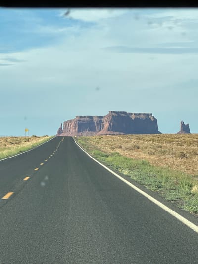 The image depicts a straight, paved road with yellow dashed lines receding into the distance. The road is flanked by dry, grassy terrain with scattered bushes and a wire fence. In the background, a large, flat-topped sandstone mesa with steep cliffs dominates the landscape. To the right of the mesa, a smaller, more slender rock formation stands. The sky is partly cloudy with a light blue hue. A yellow sign is visible on a pole to the left of the road. The scene is likely located in Oljato-Monument Valley, United States of America, known for its distinctive rock formations and desert landscape. There are no people or animals visible in the image. The overall impression is one of an open, arid expanse and a journey through a remote, scenic area.