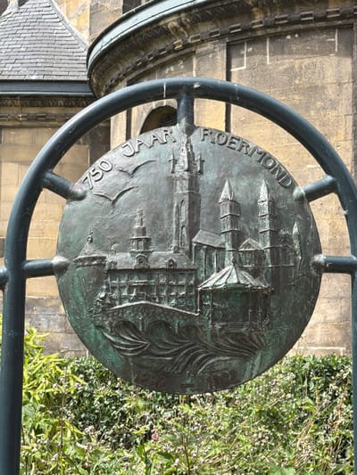 The image shows a circular bronze relief mounted on a metal frame, possibly a historical marker or artwork. The relief depicts a cityscape with buildings and spires, likely representing Roermond. Inscribed around the circumference of the relief are the words "750 JAAR ROERMOND" and the years "1232" and "1982". The relief has a green patina, indicating its age and exposure to the elements. Behind the relief, a stone building with a tiled roof is visible, suggesting an urban or historical setting. In the foreground, lush green foliage with small pink flowers partially obscures the lower portion of the relief. The scene is captured outdoors, with natural light illuminating the artwork and its surroundings. The overall impression is one of historical commemoration and artistic representation within a city.