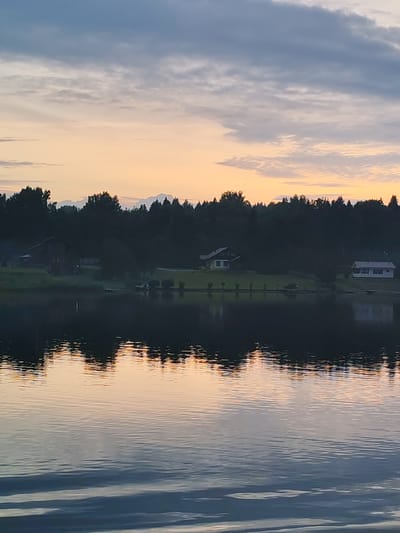 This is a vertical, eye-level, wide shot photograph of a serene lake scene at twilight, with the city of Bonema, Russia, as the implied location. The sky is filled with a mixture of soft gray and pale orange clouds, suggesting the sun has recently set or is about to rise. The water in the foreground is calm, reflecting the colors of the sky and the dark silhouette of trees and houses along the distant shore. These reflections create a mirrored image of the upper half of the scene, rippling slightly on the water's surface.
On the far shore, a dense line of dark green trees forms a backdrop. Scattered amongst the trees are a few houses, their forms indistinct in the dim light. One house, with a light-colored roof, is particularly visible on a gentle slope leading down to the water. A small, dark boat is docked near the shore. The overall impression is one of quietude and natural beauty. There are no people or animals visible in the image.