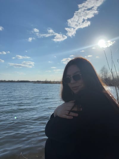 The image depicts a woman with long, dark hair and sunglasses standing near a body of water. She is wearing a dark-colored garment and holding her hand near her neck. The sky is blue with scattered white clouds and a bright sun. The water appears to be a lake or river. While the location requested is in Russia, based on the photographic evidence and without additional metadata, it is not possible to confirm that this photo was taken in Zaosinovo, Russia.