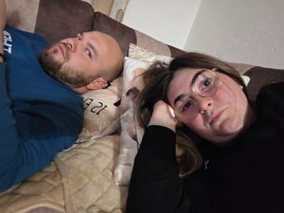 Two individuals are depicted lying side-by-side on a sofa or bed within an indoor residential environment in Negorci, North Macedonia. The male subject, positioned on the left, has a shaved head and a beard, wearing a dark teal long-sleeved shirt. He lies supine, his head resting on a light-colored pillow with a faint pattern and partially visible text, facing the camera with a neutral expression. The female subject, positioned on the right, has brown hair, wears round-rimmed glasses, and a dark long-sleeved top. She is also lying down, her left hand positioned near her head, and looks directly at the camera with a neutral expression. Both individuals are situated on a light brown, quilted blanket covering the surface. A darker brown upholstered backrest or headboard is visible behind them. The lighting is internal and appears to be artificial.