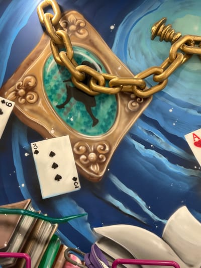This image depicts a close-up, overhead view of a mural with a three-dimensional, sculptural element of a thick, gold-colored chain. The chain is prominently featured, draped over a golden, ornate frame that encloses a silhouette of a figure falling or leaping. Inside the frame, the background is a swirling pattern of turquoise and green.
Scattered around the frame are playing cards. A card with the "9 of Spades" is visible on the left, and a "3 of Spades" is in the lower center. On the right, a portion of a card showing the "Ace of Hearts" can be seen. The background of the mural is a painted, abstract scene of rolling blue waves and white speckles that resemble stars.
In the lower left corner, a stack of books with colorful spines is partially visible, along with a bright pink railing. In the lower right, an open, white book with grey pages is depicted, and another purple object is present. The overall impression is a fantastical, dreamlike scene. The mural is located in the city of Vaganovo, Russia.