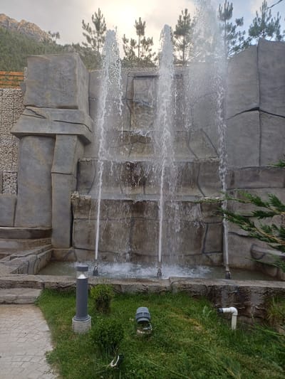 The image shows a multi-tiered artificial waterfall structure made of gray, stone-like material. Water cascades down the levels, creating a spray. In the foreground, there is a patch of green grass with several garden lights. One light is a tall, gray cylindrical lamp on a concrete base. Another, partially visible on the right, is a gray, dome-shaped light. A white pipe emerges from the ground on the right side of the frame. To the left, a paved pathway with a diamond pattern leads into the scene. In the background, pine trees are visible against a cloudy sky, with rugged, mountainous terrain in the far distance. The scene appears to be an outdoor recreational area.