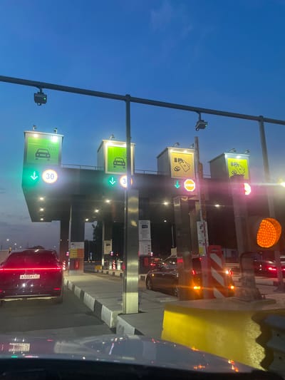 The image shows a toll plaza at dusk. The sky is a deep blue, and the lights of the plaza are illuminated. In the foreground, a dark-colored car with a license plate reading "A555KT 97" is stopped at the toll booth. The toll booth itself has signs indicating a speed limit of 30 and a green arrow, suggesting that the lane is open.

Further down the plaza, other cars are visible, some with their brake lights illuminated. There are also traffic lights and barriers. The overall scene depicts a typical toll collection point on a highway. The location is specified as the city of Shul'gino, Russia.