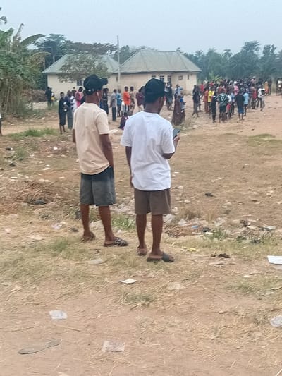 Two young men, one in a white shirt and one in a beige shirt, both wearing shorts and caps, stand facing away from the camera. Behind them is a gathering of people in an open, grassy area. A building is visible in the background, with trees to the left. Litter is scattered on the ground. The photo appears to be taken from a slightly elevated position.
