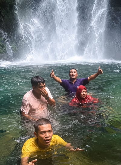 In the city of Gunung Ambat, Indonesia, a group of four individuals is captured partially submerged in the emerald-green water of a powerful waterfall. The waterfall, a cascade of white, frothy water, dominates the background. In the foreground, a man in a yellow t-shirt surfaces, his mouth open as if speaking or shouting, his left hand gesturing forward. Behind him and to his left, another man in a light pink t-shirt stands with his hand covering his nose and mouth, looking downwards. To the right, a man in a dark purple polo shirt raises both thumbs in a gesture of approval, his face turned towards the viewer. Partially visible behind him is a woman in a red headscarf and matching outfit, also in the water. The water's surface is turbulent, reflecting the dynamic energy of the waterfall.