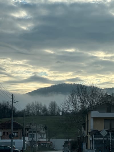 The image depicts a scene near Smilyan, Bulgaria. The sky is mostly covered with striated clouds. A mountain range runs vertically through the mid-ground, partially obscured by mist or fog. To the right, there is a mixture of vegetation, including leafless trees and grassy fields. Closer to the right edge of the image are buildings, including houses and commercial structures, along with street signs and utility poles with wires. Some vehicles are parked on the right side of the frame.