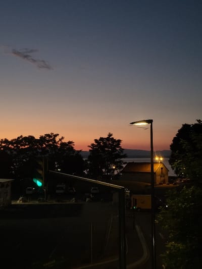 An evening shot captures a streetlamp illuminating a road and buildings under a darkening sky. The sky transitions from blue to orange near the horizon. Silhouetted trees frame the edges of the scene. A two-story structure is seen in the background, lit by its own lamps. A green traffic light is visible, indicating an active intersection.