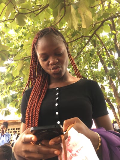 A person is facing the camera while standing outdoors with tree branches and leaves overhead. They are wearing a black top with white buttons and reddish-brown braids. Their eyes are closed, and their lips are pursed. A hand is holding a black smartphone, partially visible with a "Thank You" plastic bag and a purple garment near it. A gold bracelet is on the wrist of the hand holding the phone. In the background, a blurred figure of a person wearing blue can be seen, and the general setting appears to be a sunny, outdoor location, potentially Ezzamgbo, Nigeria, as specified.