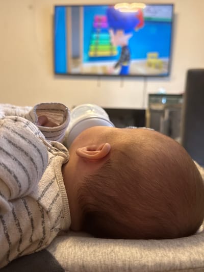 This is a close-up, eye-level shot of a sleeping infant, positioned on its side. The infant's head, seen from the back and right profile, is the primary focus, with fine, dark hair visible. A baby bottle, partially obscured by the infant's head, rests nearby. The infant is dressed in a light-colored garment with a striped pattern on the sleeves, and a grey blanket is visible underneath. In the blurred background, a television screen displays a cartoon featuring animated characters. The scene appears to be set in an indoor domestic environment, possibly a living room, with a wall and furniture suggested in the background. There is no specific indication of the city of Oldham, United Kingdom, within the visual information provided in the image.