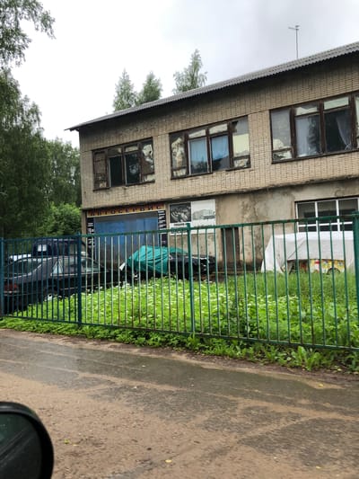 An overcast, daytime exterior shot shows a two-story, light-colored brick building with a corrugated metal roof, located in Safonovo, Russia. The building appears to be an auto service center, with large blue garage doors and signage visible on the facade. In front of the building, a green metal fence encloses a grassy area with several parked cars. One car is covered with a green tarp, and another is partially obscured by a white tarpaulin. The ground in front of the fence is a wet, asphalt road, suggesting recent rain. Trees surround the property, and the sky is a uniform grey.